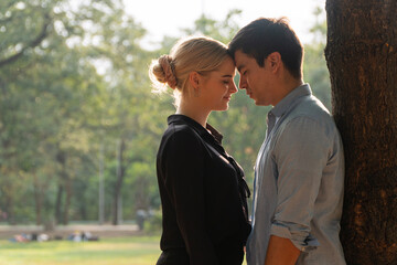 Fototapeta premium Romantic young Caucasian couple standing close with foreheads touching and eyes closed under a tree in a green park. Beautiful sunlight. Perfect for love, romance, and outdoor lifestyle concepts.
