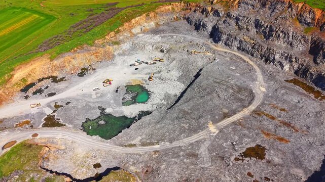 Elevated view of deep stone excavation site with machinery, trucks and emerald green water pools. Open pit industrial rock quarry, Burgas region, Bulgaria