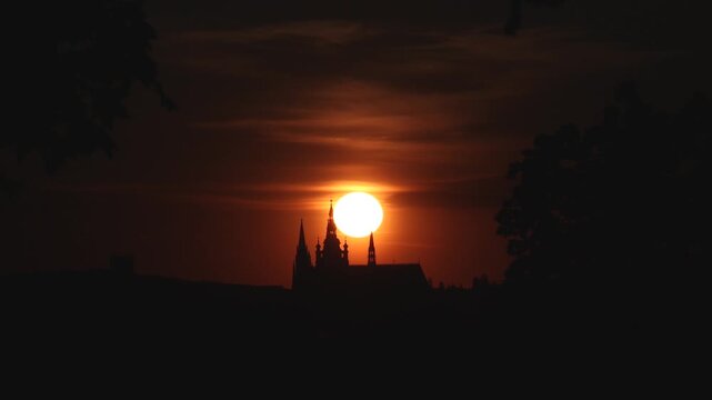 Sunset glows behind darkened church spires of Prague Castle. Trees frame the silhouette against orange sky in beautiful Czech scene.