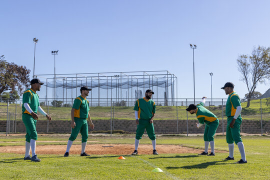Five male players in green-yellow jerseys drilling near mound with orange cones, batting cage