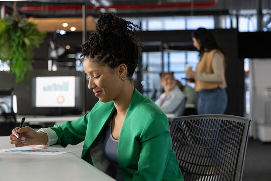 African American woman wearing green blazer writing on document at open-plan office desk with pen