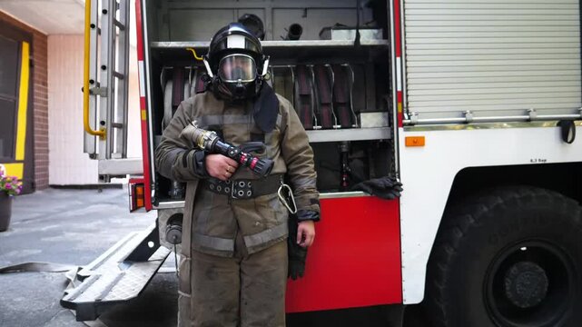 Portrait of young fireman in full equipment standing near a fire engine. Male firefighter with fire hose in hand looking into camera against the background of a big red car. Concept of saving lives