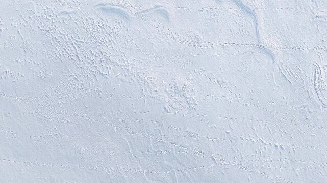 Drone video captures a pristine snowfield from above, revealing intricate wind-carved patterns and faint tracks under soft natural daylight. No people or wildlife in view. 199 characters.