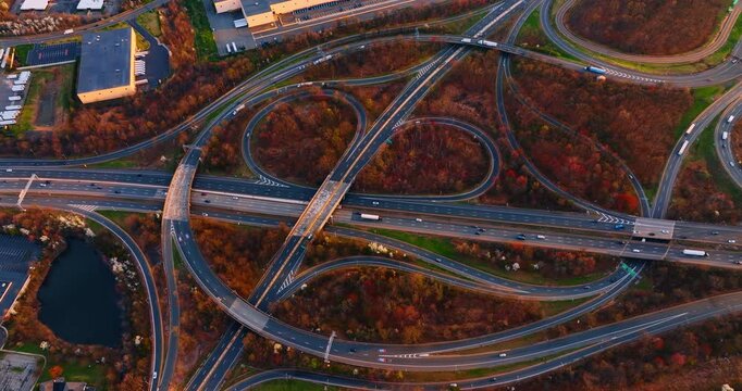 Cloverleaf highway interchange surrounded by autumn woods, New Jersey. Top-down of looping cloverleaf ramps and an overpass weaving through colorful seasonal trees and warehouses
