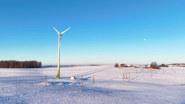 Drone video captures a solitary wind turbine amid vast snow-covered fields, crisp winter air, and vibrant blue sky, with distant country buildings and long dramatic shadows.