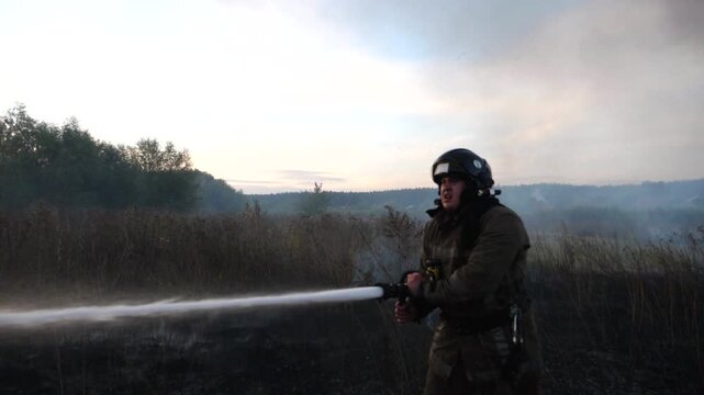 Male firefighter in uniform extinguishes burning dry grass using fire hose at countryside. Professional fireman in full equipment putting out flame on field. Heroic profession and fire safety concept