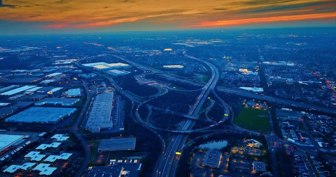 Aerial view of a freeway interchange in blue twilight, New Jersey. High overhead of intertwining ramps and lanes in cool dusk tones with warehouses spreading to a fading horizon