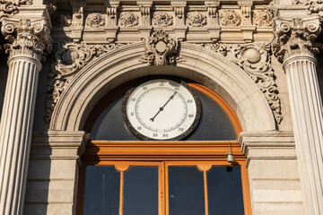Historic stone facade with an ornate round barometer above an arched wooden window, flanked by Corinthian columns and decorative sculpted details, conveying heritage, time and classical architecture © evannovostro