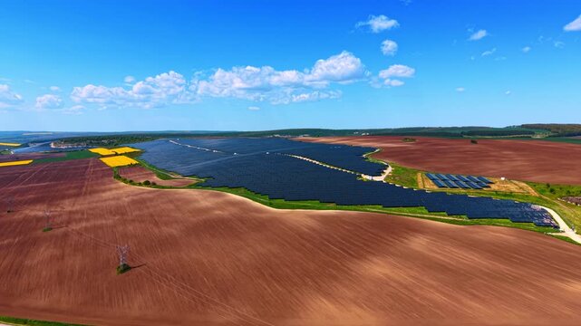 Panoramic photovoltaic farm across plowed fields under blue sky. Wide overhead sweep showing dark panel arrays bordering bare brown soil with pylons and distant tree line.