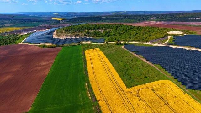 Solar panels beside rapeseed and farmland fields from above. Birds eye composition of photovoltaic rows next to plowed soil, green crop and flowering yellow rapeseed strips.
