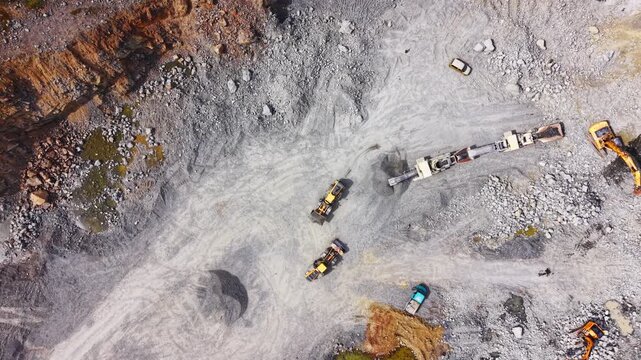 Top down drone view of multiple excavators and mobile crushing units operating on the rocky pit floor. Overhead shot of heavy machinery in a stone quarry.