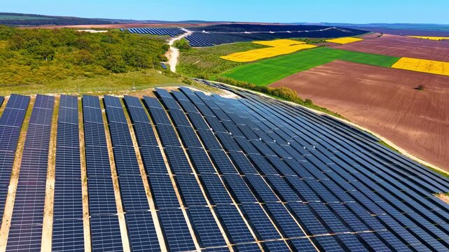 Tilted solar modules on slope with patchwork crops beyond. Aerial along banked panel ranks descending toward a quilt of brown, green and yellow farm plots.