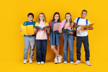 Obraz premium Happy school children with backpacks and books posing confidently on yellow studio background, representing friendship, education and modern student lifestyle
