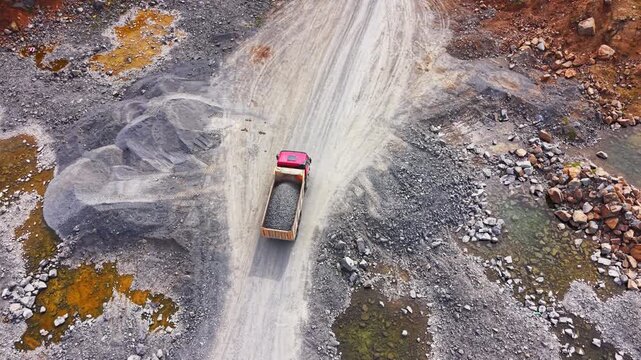 Overhead view of a red tipper commercial vehicle driving on a gravel road past muddy puddles. Dump truck transporting crushed stone through a mine