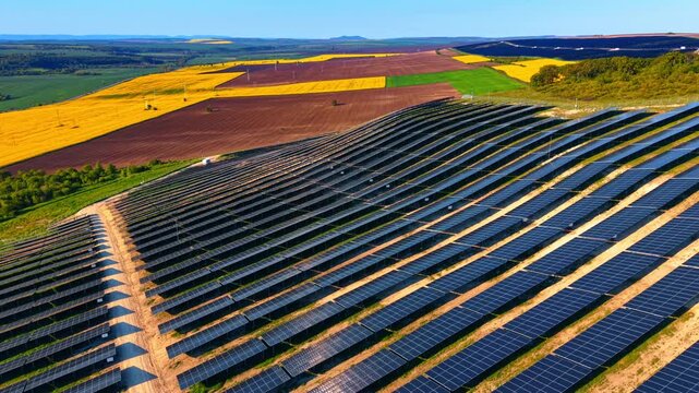 Sloped solar farm overlooking rapeseed and plowed crop fields. Aerial of banked module rows above a patchwork of canola, bare soil and green plots stretching to hills.