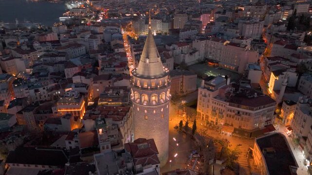 Morning view of Galata Tower illuminated against historic Golden Horn and cityscape in Istanbul with lights illuminating streets and buildings at sunrise. Flying around iconic Galata Tower in sunset.