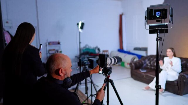 Two operators filming woman in white suit on studio sofa. Bald cameraman and assistant with phone work telephoto rigs as a seated subject gestures on a leather couch.