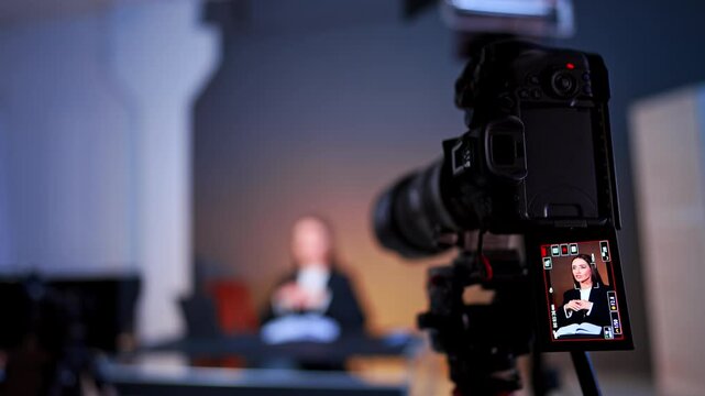 Camera viewfinder framing woman during studio interview shoot. Close shot of a screen displaying a seated interviewee while the subject sits blurred in warm background.