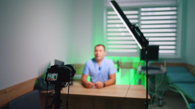 Studio camera filming man at desk under green ambient lighting. Foreground rig with rear preview and a blurred presenter seated behind a wooden desk near blinds.