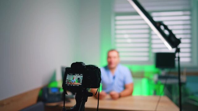 Mirrorless camera filming presenter seated at desk with green light. Rig in focus with a blurred speaker behind, settings visible on the rear display under soft daylight.