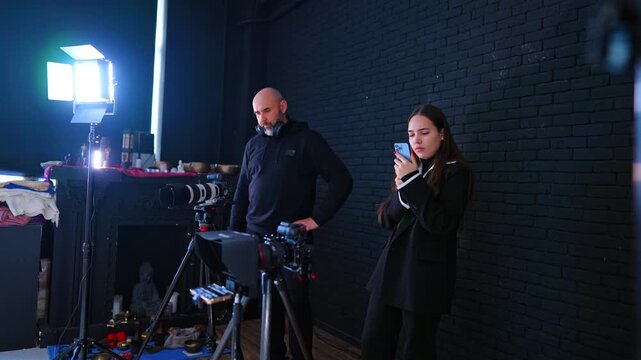 Film crew filming interview in dark studio with cinema light. Bearded operator and woman reviewing a phone beside cameras on tripods against a black brick backdrop.