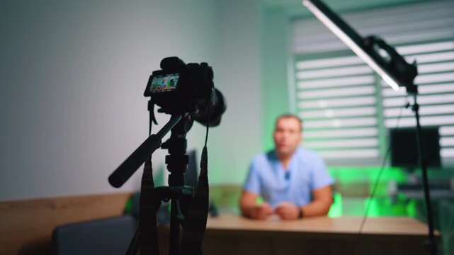 Camera and boom filming seated presenter in green-toned office. Rig with strap and microphone in focus, blurred speaker in scrubs behind under window blinds.