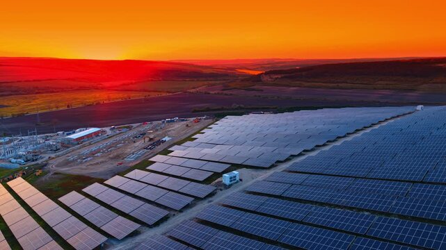Solar power plant under construction at vivid orange sunset. Dusk aerial of panel rows beside a substation, site cabins and machinery against a fiery sky.
