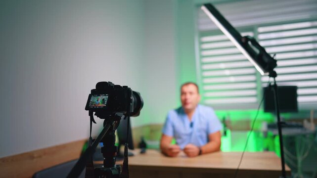 Camera on tripod recording man at desk in green-lit office. Foreground rig with rear screen shows a blurred presenter seated by a window with venetian blinds.
