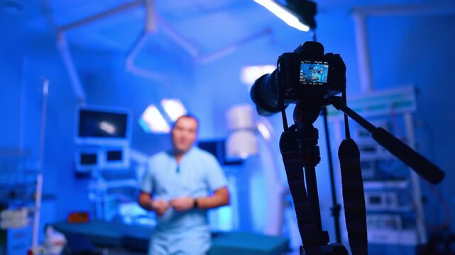 Camera filming doctor standing in blue surgical operating room. Tripod rig with rear preview shows a blurred presenter in scrubs amid surgical lamps and monitors.