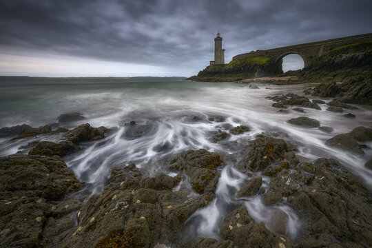 View of Phare du Petit Minou lighthouse and its stone bridge with waves crashing against the rocky shore under a dramatic cloudy sky Brittany, France.