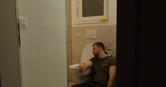 A man sits on the bathroom floor beside a toilet, looking unwell and nauseous, representing sickness, stomach pain, food poisoning, hangover, illness, exhaustion, discomfort