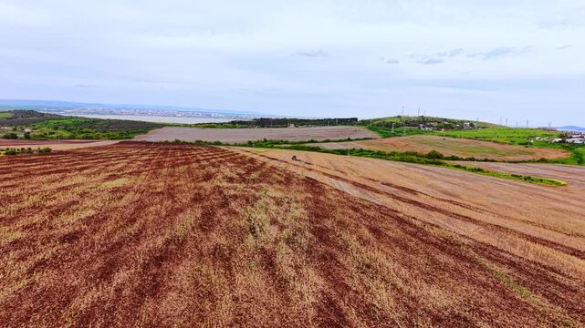 Red-brown soil rows converge toward horizon with distant lake and city skyline under overcast sky. Tractor cultivating rolling agricultural fields near Varna, Bulgaria