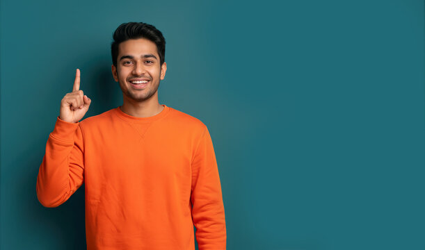 Young man smiling, pointing index finger up, plain teal background with copy space, excellent for showing ideas or suggestions.