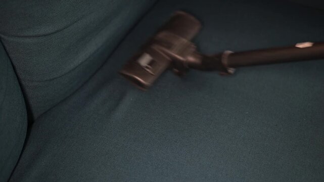 A person uses a vacuum cleaner to clean a couch in a living room. The action focuses on removing dirt and debris from the fabric surface to keep the area tidy