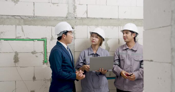 Asian male manager gestures beside female engineer using laptop and worker holding tool, leading construction document review process and safety coordination meeting indoors with project team.