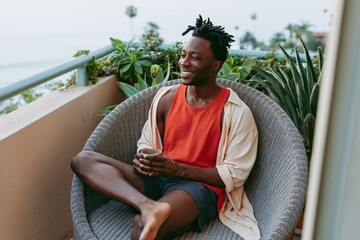 Fototapeta premium Smiling man seated in wicker balcony chair holding coffee among plants for lifestyle banner and background design