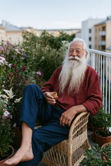Fototapeta premium Smiling senior man with long white beard on a balcony holding coffee in a wicker chair for cozy outdoor lifestyle and garden portrait