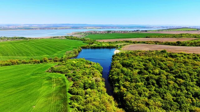 Elevated panoramic frame, foreground wheat field slopes to tree-lined river, blue lake extends to middle ground with village and distant sea on horizon.