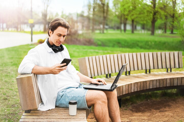 Obraz premium Freelancer doing remote work on a laptop outdoors. Man sitting on a park bench checking his smartphone with a coffee cup nearby.
