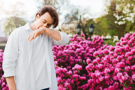 Sick man outdoors suffering from spring allergy and hayfever. He is sneezing into his elbow due to pollen from the bright pink blooms and flowers.