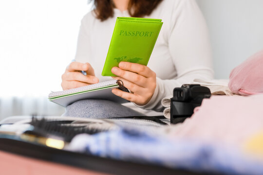Woman writing a packing list and holding a green passport for travel preparation. She is sitting by an open suitcase before a summer vacation.