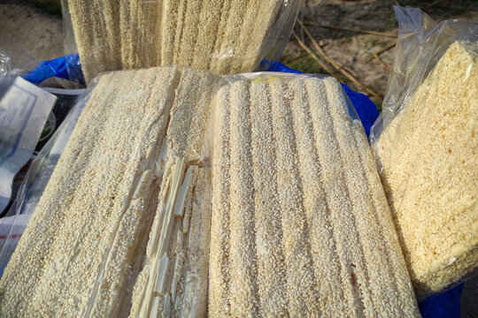 Freshly prepared, delicious sesame seed khaja sweets are stacked in transparent packaging at an outdoor rural market. The popular traditional Asian confectionery with rich texture and crispy layers.