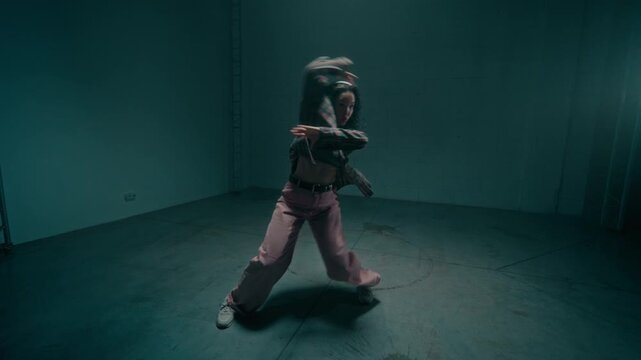 Cinematic zoom out shot of young woman putting on headphones and performing freestyle hip hop choreography in empty underground studio lit by flashing lights, copy space