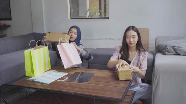 Two friends unpacking colorful shopping bags on livingroom sofa, woman with hijab and companion compare purchases and open small