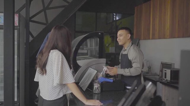 Asian barista hands receipt to customer at modern cafe counter, warm friendly exchange with male staff in apron smiling