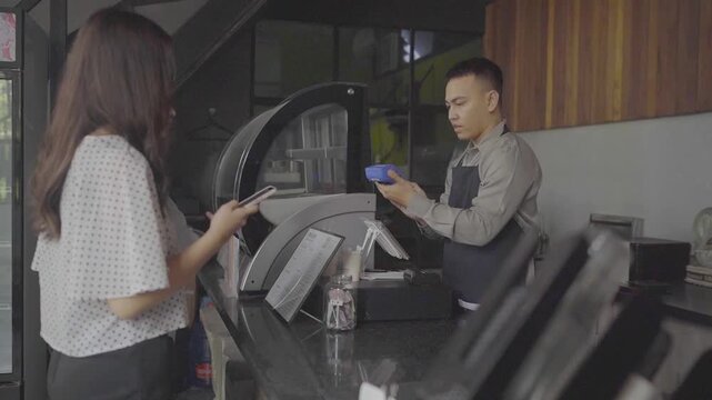 Asian cashier female customer mobile payment at cafe counter, male barista in apron uses blue card reader while woman presents