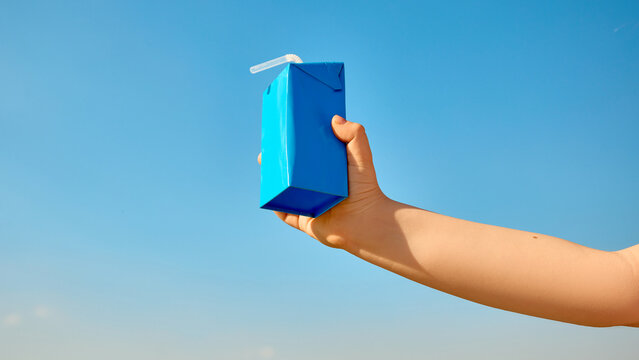 Child hand holding blue drink carton with straw against clear sky. Concept of kids beverage packaging, school lunch, dairy drink, juice box advertising, summer snack, and label mockup.