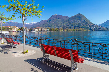 Lugano, Switzerland, a red bench on a sunny lakeside promenade overlooking calm blue water and forested mountains under a clear sky © dancar