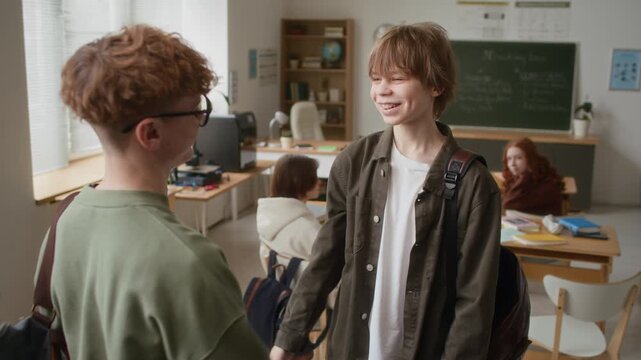 Teenage boy with curly hair and glasses falling for fake handshake prank, pulled by his friend in classroom