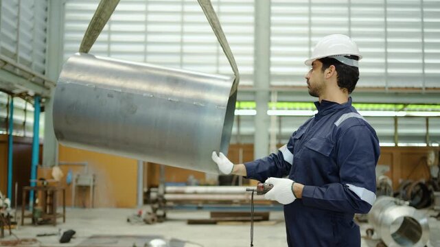 An industrial engineer wearing a hardhat and uniform operates an overhead crane remote control to lower a heavy metal pipe cylinder inside a manufacturing factory. Professional machine operator.